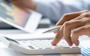 A close-up of an accountant's hand using a calculator with financial documents and charts, highlighting the importance of accountant insurance and bookkeeper insurance. Huff Insurance in Pasadena, Maryland offers comprehensive coverage to protect your career.