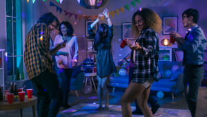 Diverse group of teenagers enjoying a graduation party at home, dancing and holding red cups, in a lively room decorated with colorful lights and streamers. Image emphasizes the importance of understanding graduation party risks, as discussed by Huff Insurance.